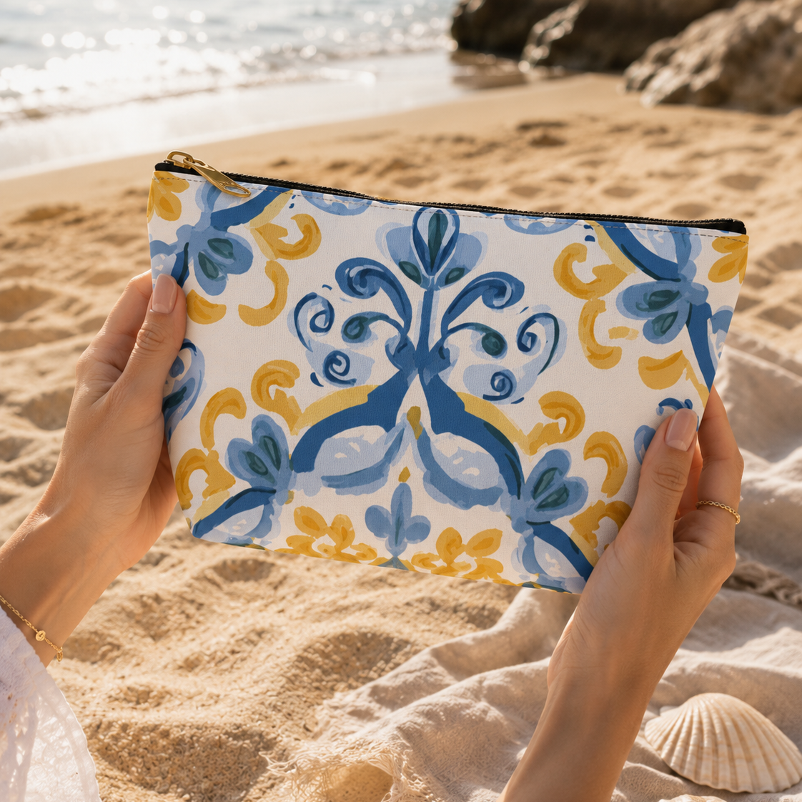 Person holding a patterned pouch on a sandy beach with rocks and water in the background