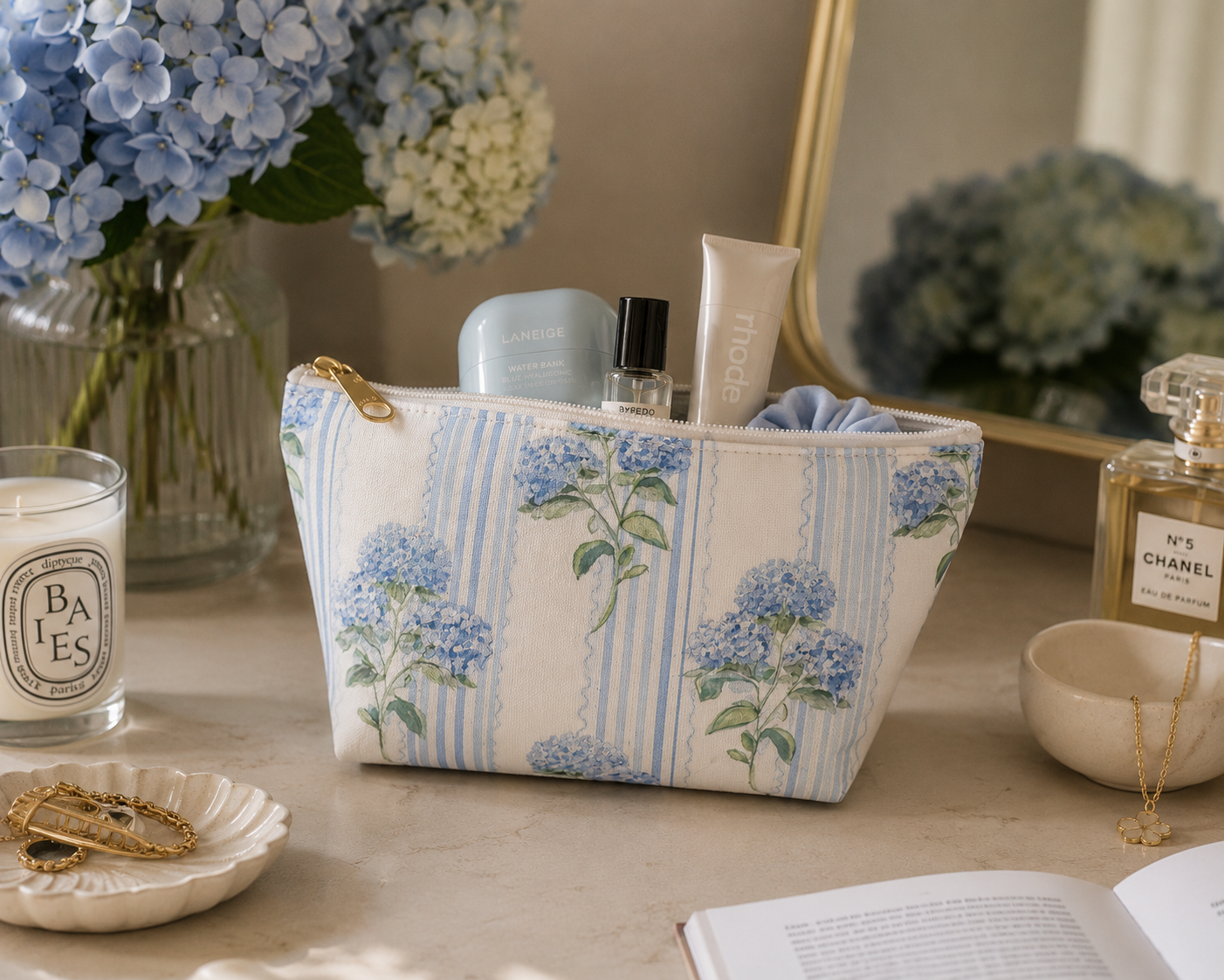 Floral-patterned hydrangea cosmetic bag on a table with candles, books, and blue hydrangeas in the background