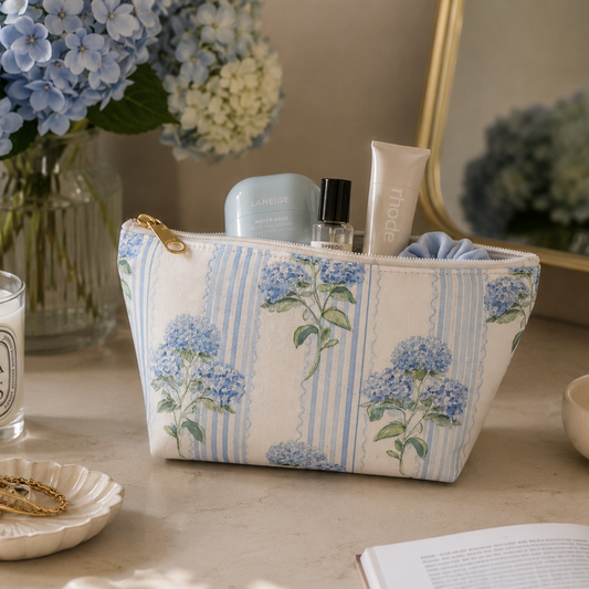 Floral-patterned hydrangea cosmetic bag on a table with candles, books, and blue hydrangeas in the background