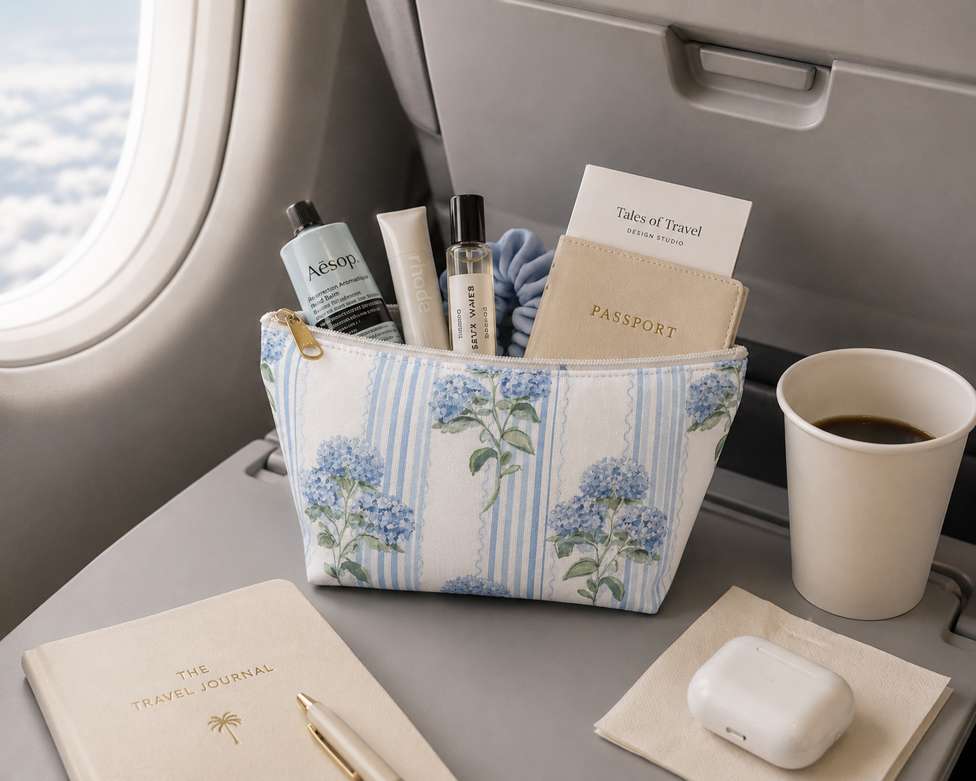 Travel essentials including a passport, coffee, and a floral-patterned pouch with hydrangeas on an airplane tray table.