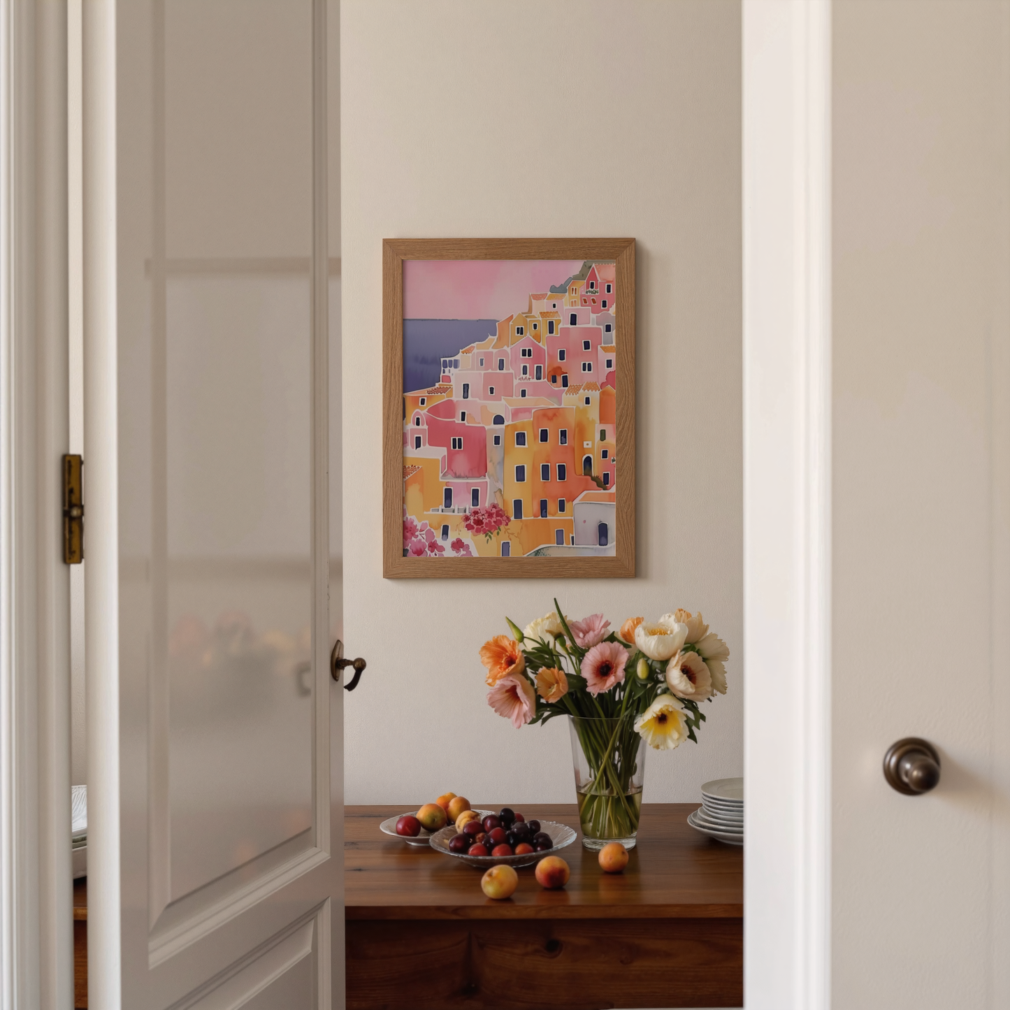 Framed artwork of colorful buildings on a wall above a table with flowers and fruit.
