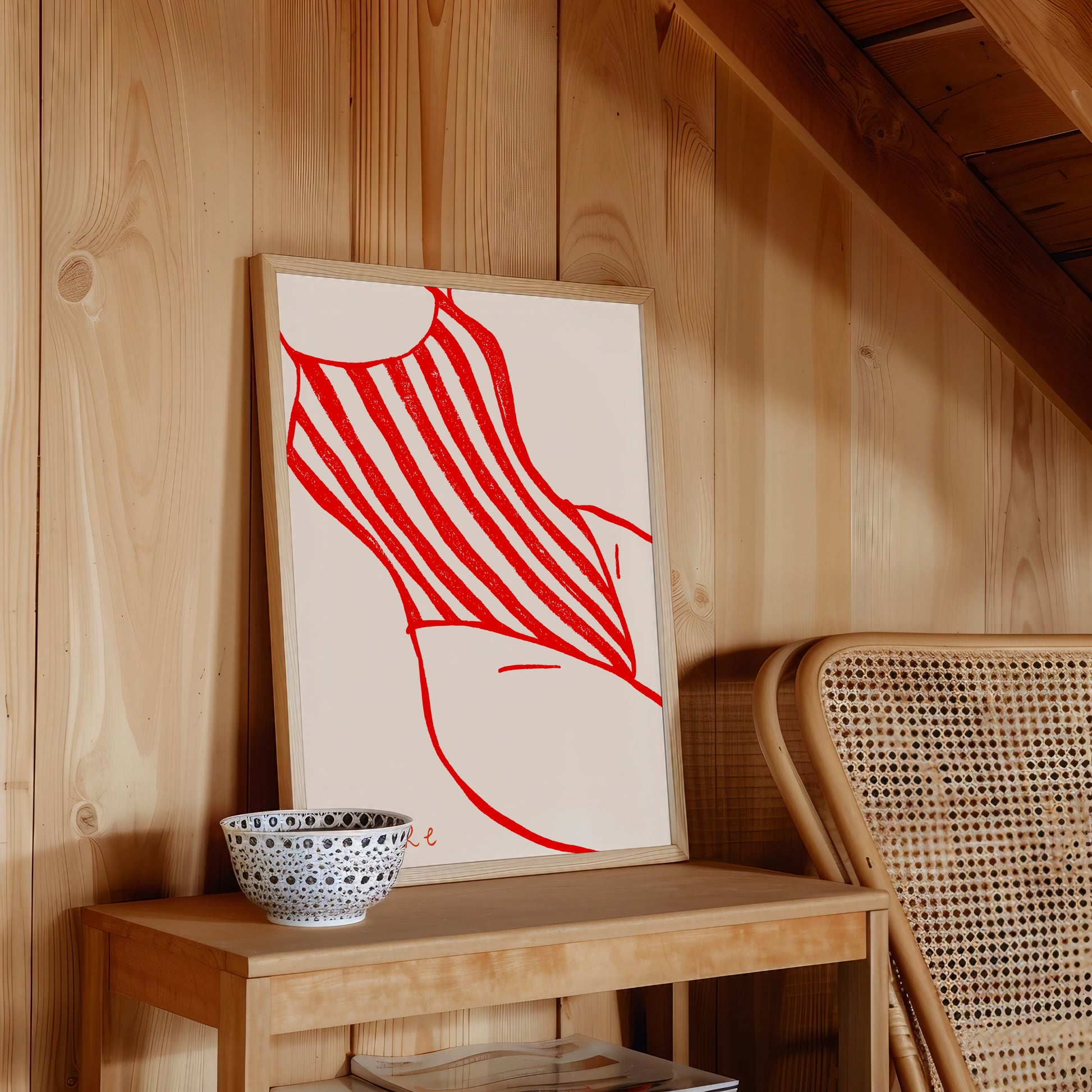 Framed artwork with a red abstract female body with swimsuit design on a wooden surface next to a wicker chair.