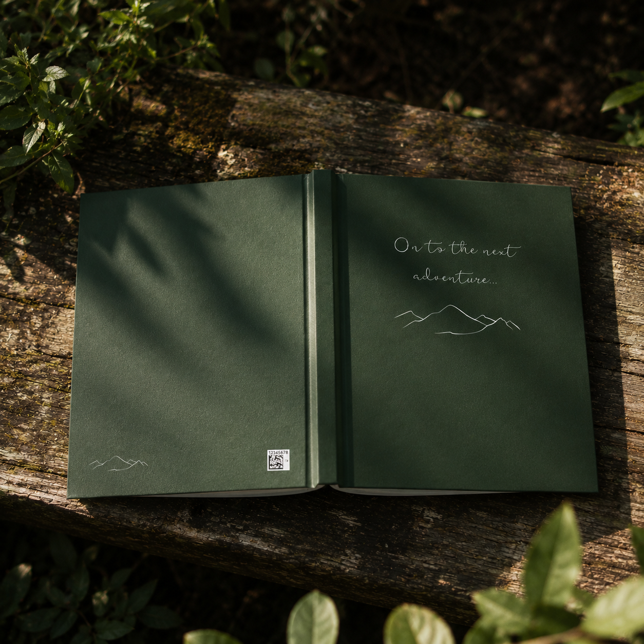 Open green notebook with 'On to the next adventure' text and mountain illustration on a wooden surface with plants.