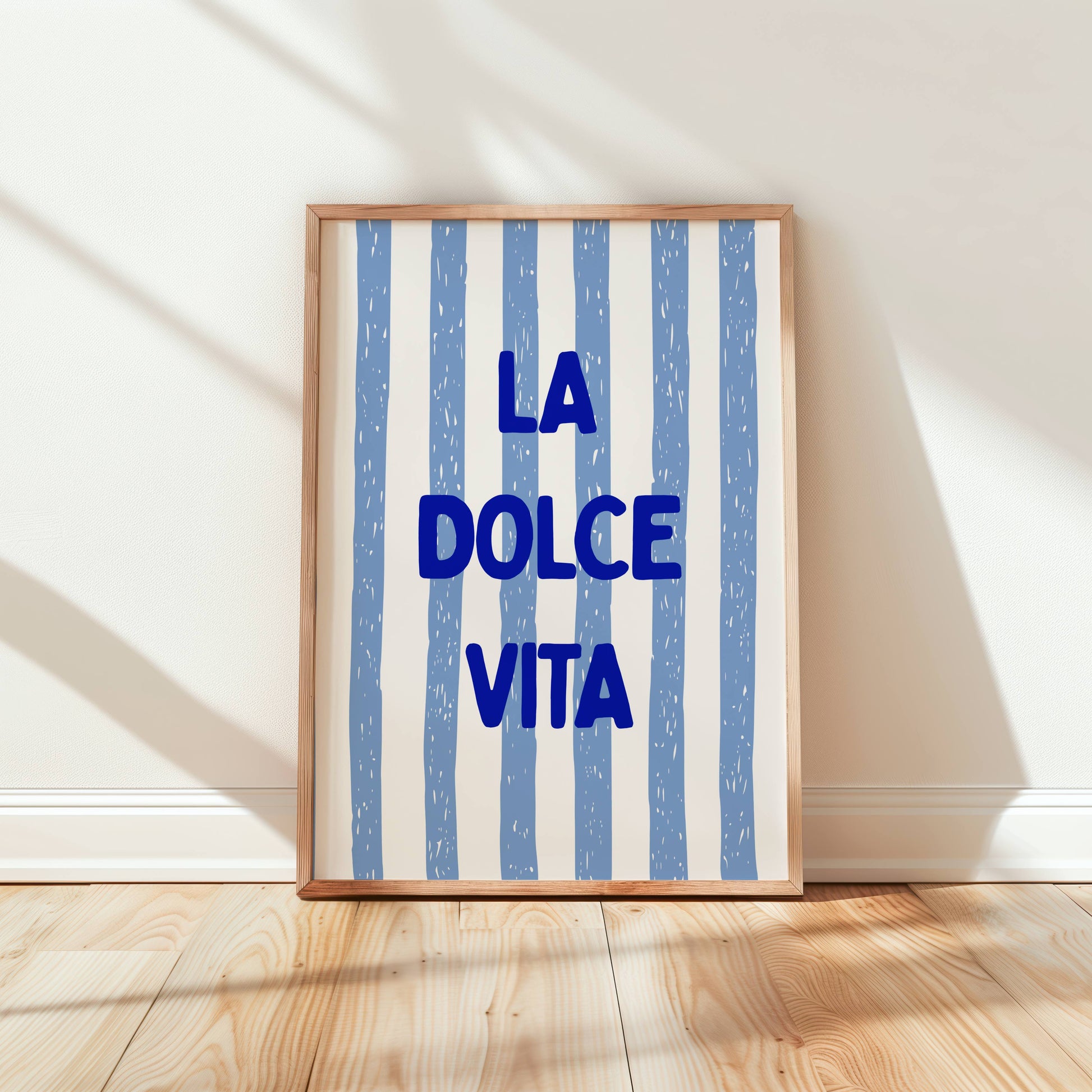 Framed artwork with 'La Dolce Vita' text on a striped background in a room with wooden floor and white walls.