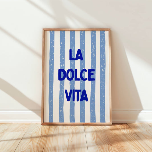 Framed artwork with 'La Dolce Vita' text on a striped background in a room with wooden floor and white walls.