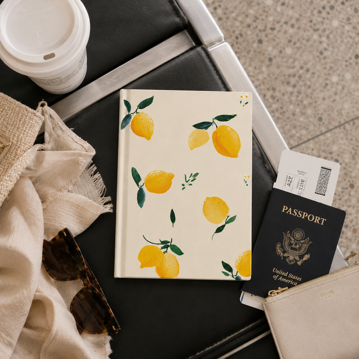 Notebook with lemon design, passport, sunglasses, and coffee cup on a suitcase.