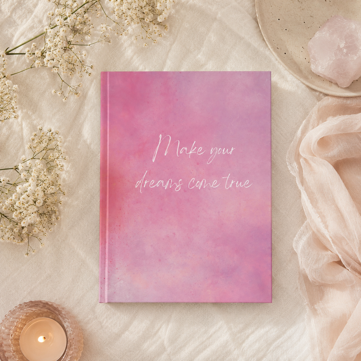 Pink watercolor notebook with 'Make your dreams come true' text, surrounded by flowers and a candle on a light surface.