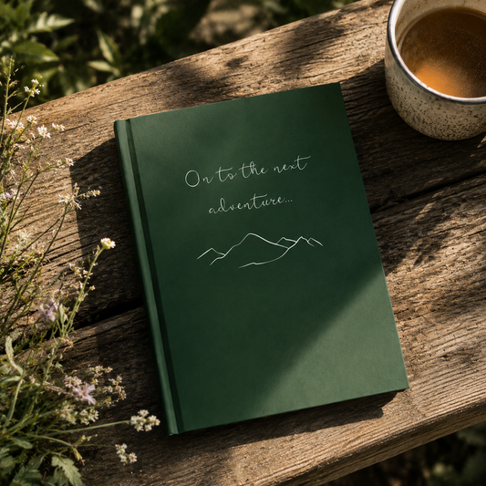 Green notebook with 'On to the next adventure' text on a wooden surface with a cup of coffee and flowers.