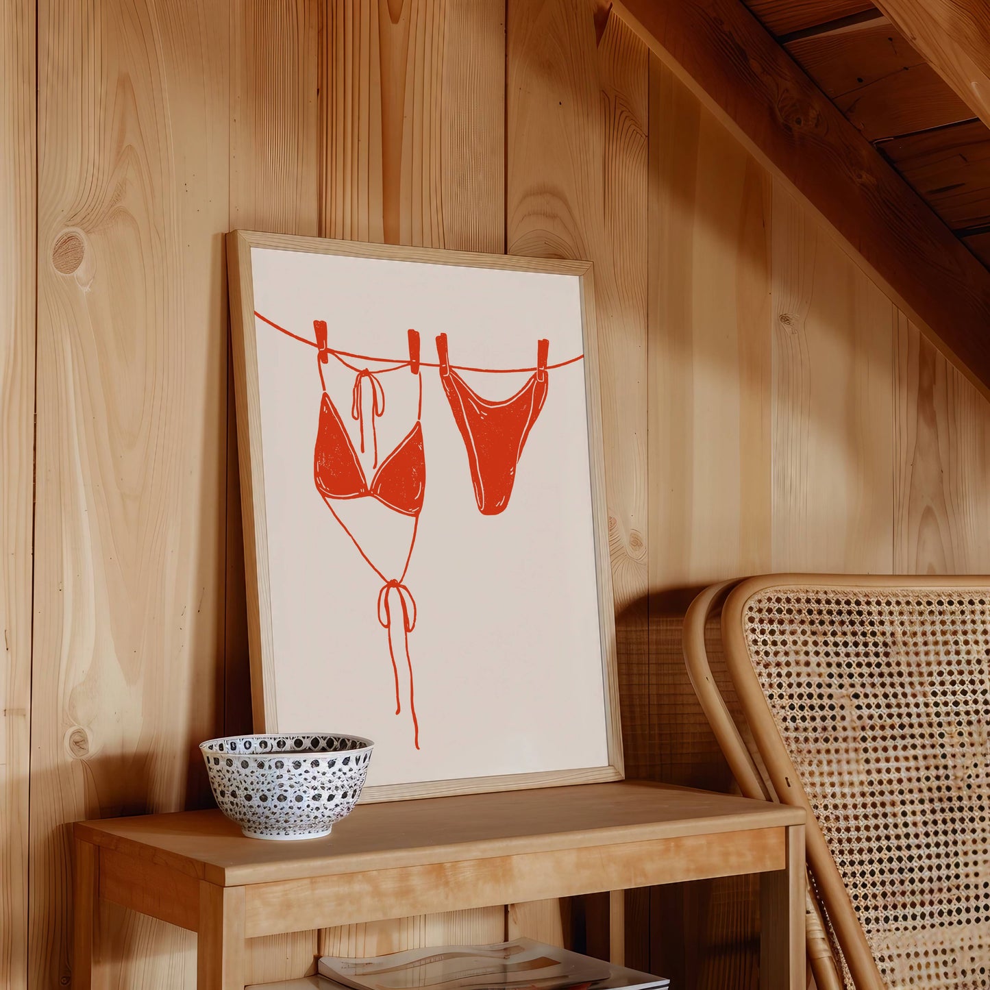 Framed artwork of a red bikini on a clothesline above a wooden table with a bowl and chair.